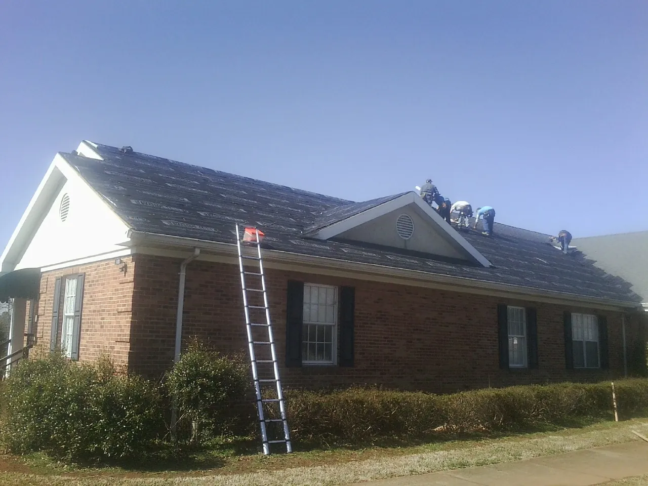 Crew tearing shingles off a brick ranch roof with a ladder set — Emergency Roof Repair work by our crew in Colonia