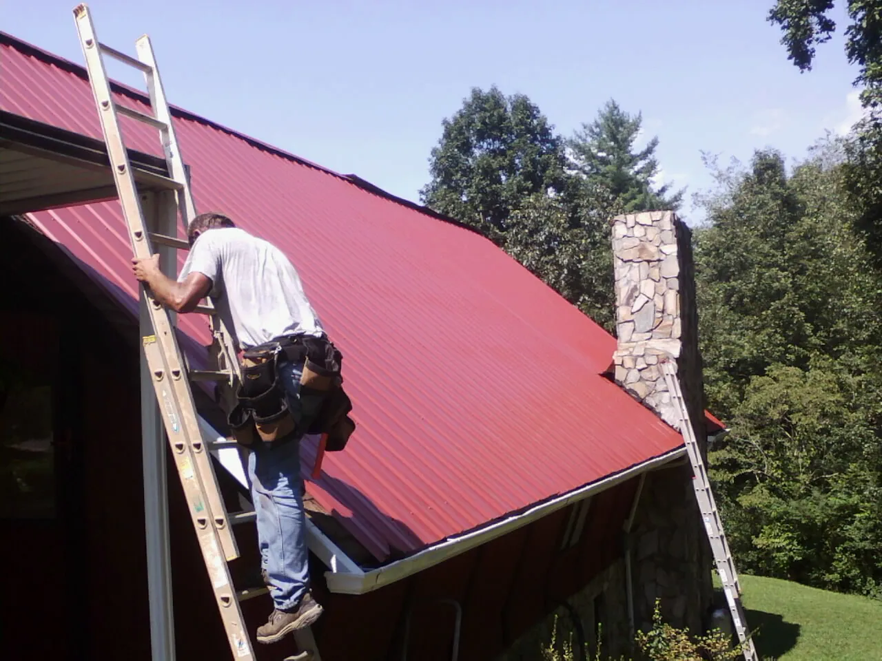 Roofer on a ladder beside a red metal roof and stone chimney — Soffit & Fascia Repair by Roofing Brothers in Colonia